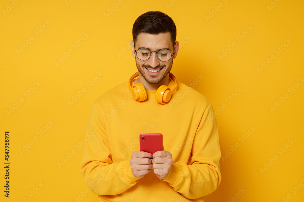 Hipster guy in bright sweatshirt wearing headphones on neck, typing phone messages to friends with happy smile, isolated on yellow background