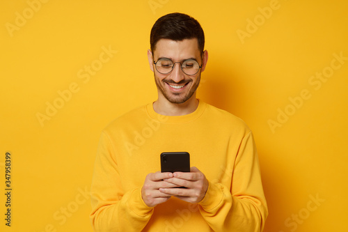 Young man looking at mobile phone screen through glasses, laughing at jokes sent by friends, dressed in bright clothes, isolated on yellow background