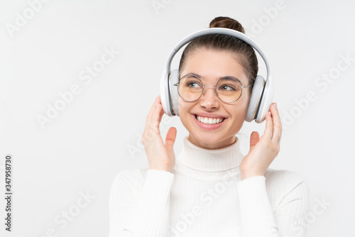 Young girl in eyewear and white turtleneck sweater, listening to favorite music via wireless headphones, smiling happily, feeling relaxed, isolated on gray background