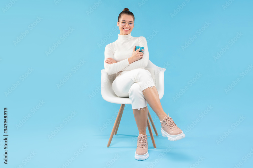 Young trendy positive girl sitting in armchair, holding coffee cup in hands, enjoying her free time from work, isolated on blue background Young trendy positive girl sitting in armchair, holding coffee cup in hands, enjoying her free time from work, isolated on blue background