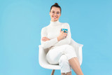 Young smiling woman sitting in white armchair in relaxed pose drinking tea from cup, keeping legs and arms crossed to feel comfortable, isolated on blue background
