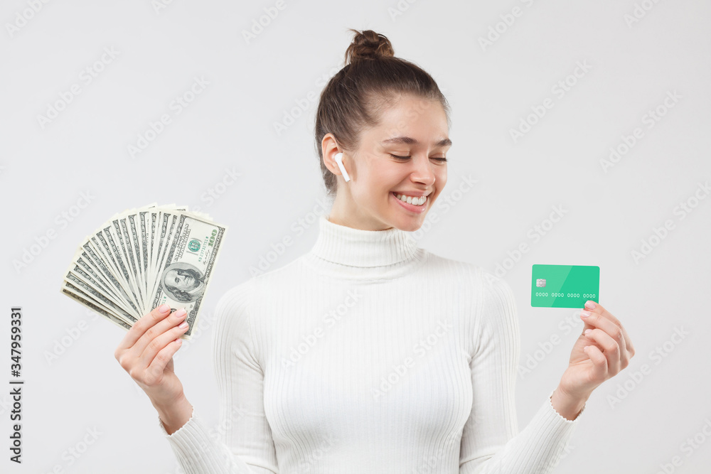 Young woman in white turtleneck, choosing between using dollars in cash or money on credit card, isolated on gray background Young woman in white turtleneck, choosing between using dollars in cash or money on credit card, isolated on gray background