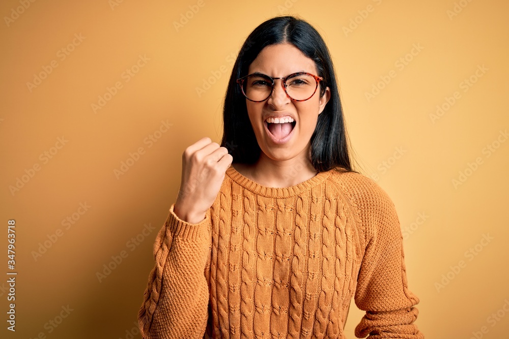 Young beautiful hispanic woman wearing glasses over yellow isolated background angry and mad raising fist frustrated and furious while shouting with anger. Rage and aggressive concept. Young beautiful hispanic woman wearing glasses over yellow isolated background angry and mad raising fist frustrated and furious while shouting with anger. Rage and aggressive concept.