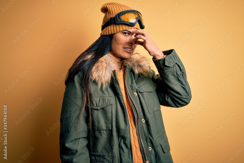 Young beautiful hispanic woman wearing ski glasses and coat for winter weather smelling something stinky and disgusting, intolerable smell, holding breath with fingers on nose. Bad smell