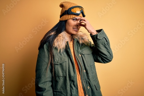 Young beautiful hispanic woman wearing ski glasses and coat for winter weather smelling something stinky and disgusting, intolerable smell, holding breath with fingers on nose. Bad smell © Krakenimages.com