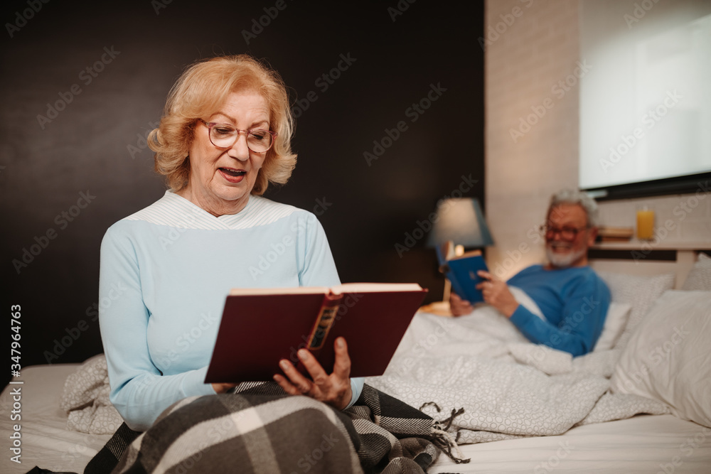 Φωτογραφία Portrait of smiling blonde woman sitting on bed and reading book to her husband who is lying in bed behind her