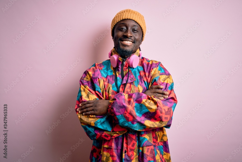 Young handsome african american man wearing colorful coat and cap over pink background happy face smiling with crossed arms looking at the camera. Positive person.