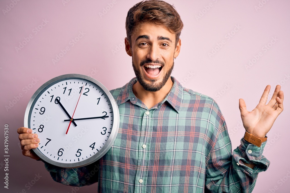 Young man with beard doing countdown using big clock over isolated pink background very happy and excited, winner expression celebrating victory screaming with big smile and raised hands Young man with beard doing countdown using big clock over isolated pink background very happy and excited, winner expression celebrating victory screaming with big smile and raised hands
