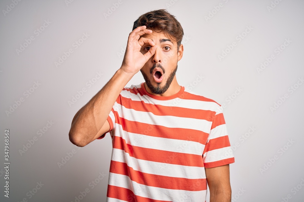 Young handsome man with beard wearing striped t-shirt standing over white background doing ok gesture shocked with surprised face, eye looking through fingers. Unbelieving expression. Young handsome man with beard wearing striped t-shirt standing over white background doing ok gesture shocked with surprised face, eye looking through fingers. Unbelieving expression.