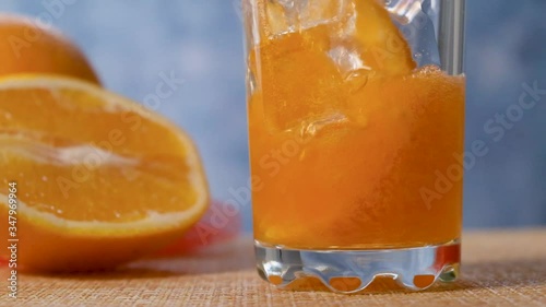 Yellow orange lemonade is poured into a clear glass with orange and ice. Slow motion.
