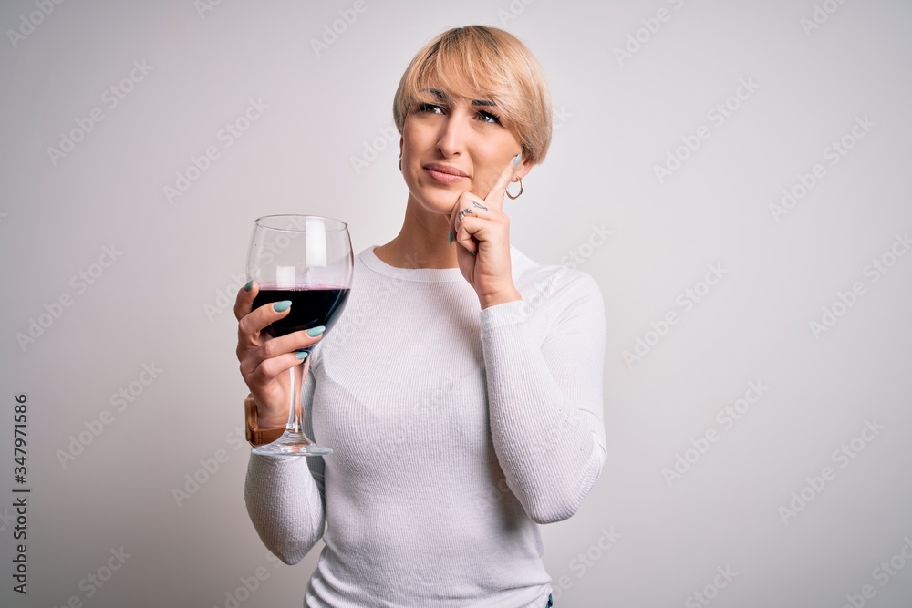 Young blonde woman with short hair drinking a glass of red wine over isolated background serious face thinking about question, very confused idea Young blonde woman with short hair drinking a glass of red wine over isolated background serious face thinking about question, very confused idea