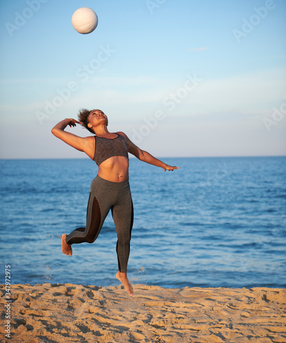 Stunning young woman serving volleyball at sunset on a beach with ocean behind her