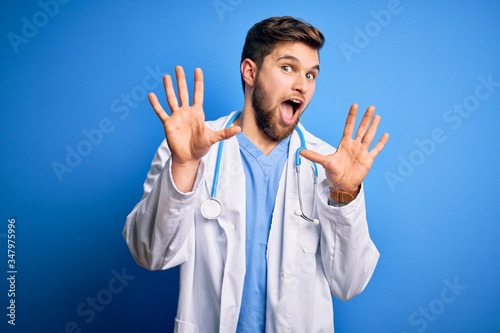 Young blond doctor man with beard and blue eyes wearing white coat and stethoscope afraid and terrified with fear expression stop gesture with hands, shouting in shock. Panic concept.