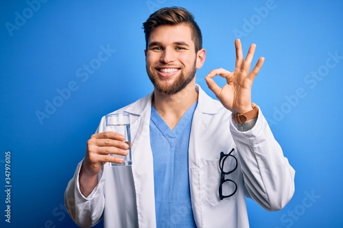 Young blond doctor man with beard and blue eyes wearing coat drinking glass of water doing ok sign with fingers, excellent symbol