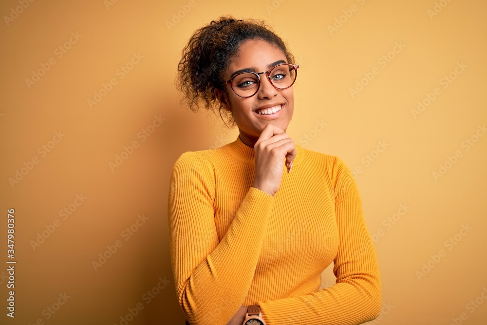 Young beautiful african american girl wearing sweater and glasses over yellow background looking confident at the camera smiling with crossed arms and hand raised on chin. Thinking positive. Young beautiful african american girl wearing sweater and glasses over yellow background looking confident at the camera smiling with crossed arms and hand raised on chin. Thinking positive.