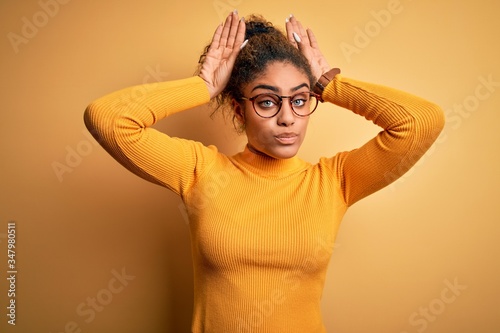 Young beautiful african american girl wearing sweater and glasses over yellow background Doing bunny ears gesture with hands palms looking cynical and skeptical. Easter rabbit concept. photo