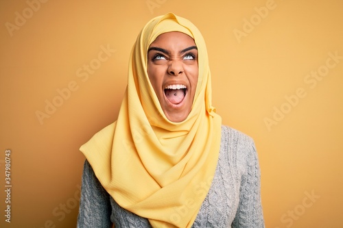 Young beautiful african american girl wearing muslim hijab over isolated yellow background angry and mad screaming frustrated and furious, shouting with anger. Rage and aggressive concept. © Krakenimages.com