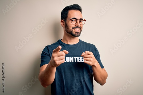 Handsome man with beard wearing t-shirt with volunteer message over white background pointing fingers to camera with happy and funny face. Good energy and vibes.