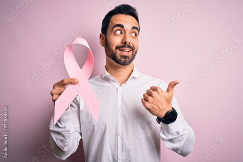 Young handsome man with beard holding pink cancer ribbon over isolated background pointing and showing with thumb up to the side with happy face smiling © Krakenimages.com