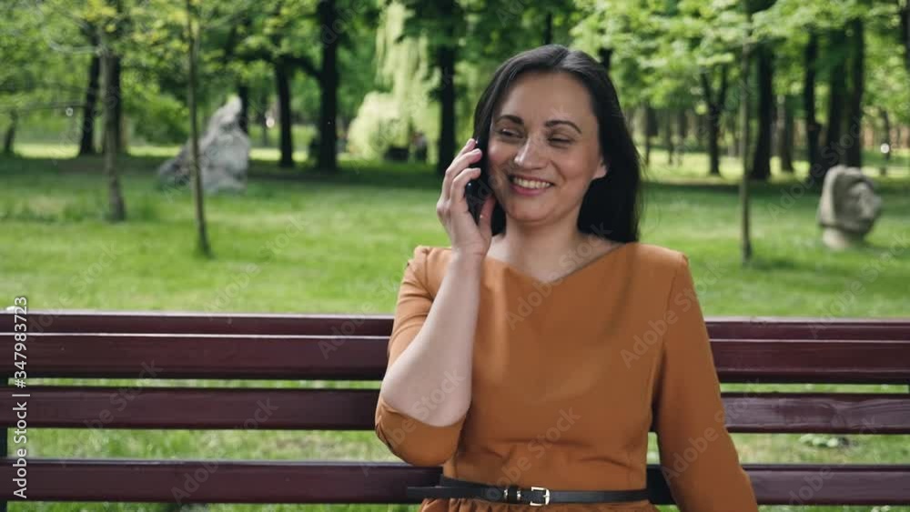 A young brunette sits on a bench in a park. Beautiful woman speaks on the phone and smiles. A young brunette sits on a bench in a park. Beautiful woman speaks on the phone and smiles.
