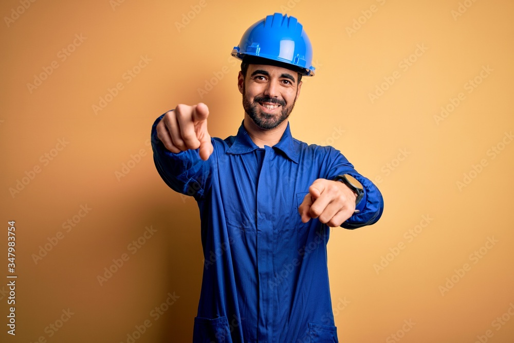 Mechanic man with beard wearing blue uniform and safety helmet over yellow background pointing to you and the camera with fingers, smiling positive and cheerful