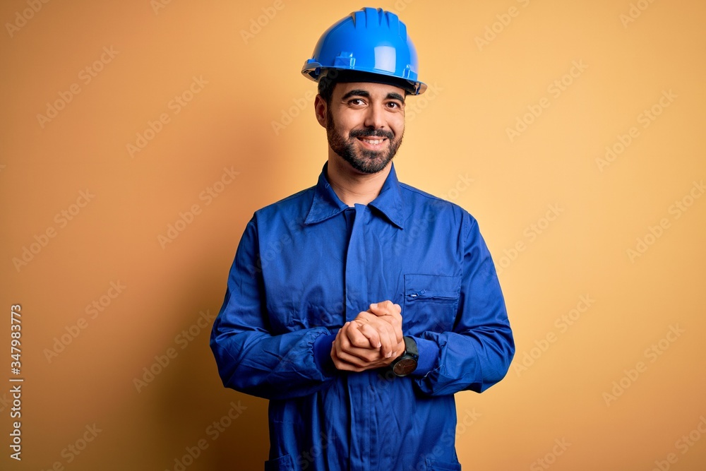 Mechanic man with beard wearing blue uniform and safety helmet over yellow background with hands together and crossed fingers smiling relaxed and cheerful. Success and optimistic