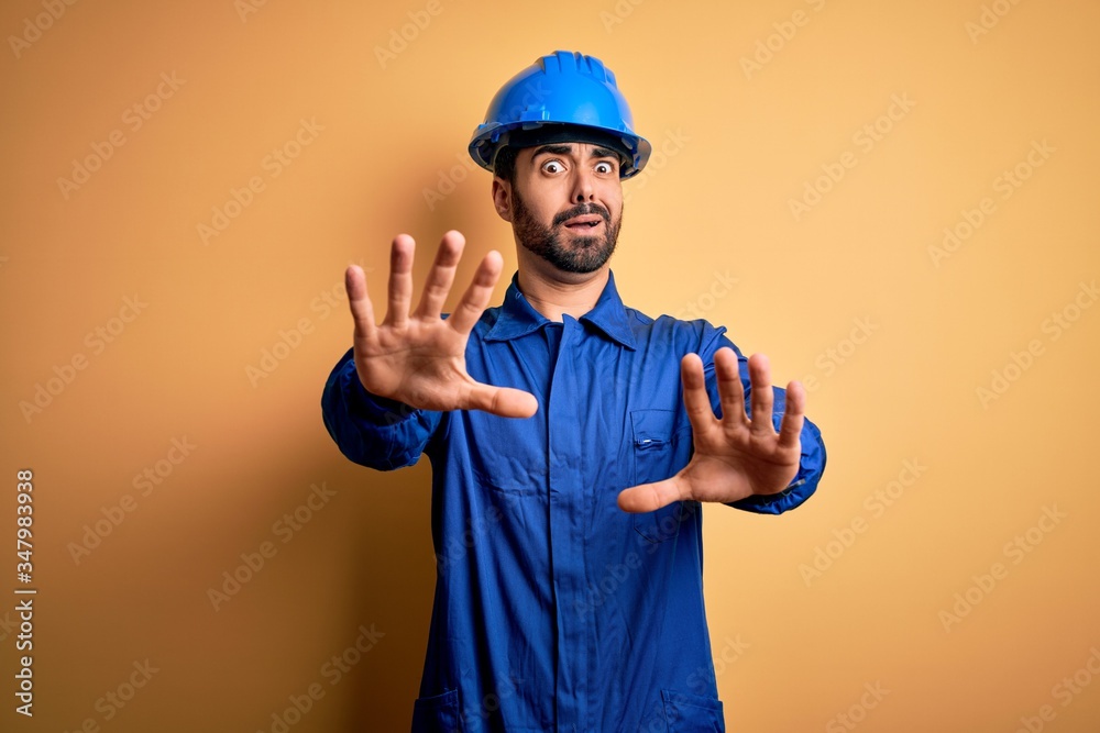 Mechanic man with beard wearing blue uniform and safety helmet over yellow background afraid and terrified with fear expression stop gesture with hands, shouting in shock. Panic concept.