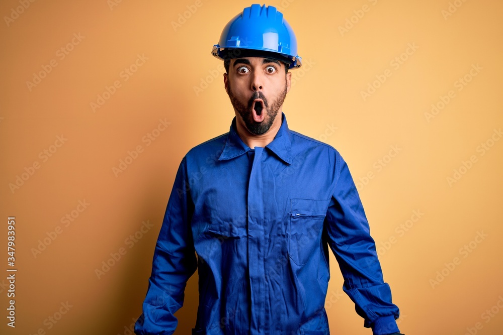 Mechanic man with beard wearing blue uniform and safety helmet over yellow background afraid and shocked with surprise and amazed expression, fear and excited face.