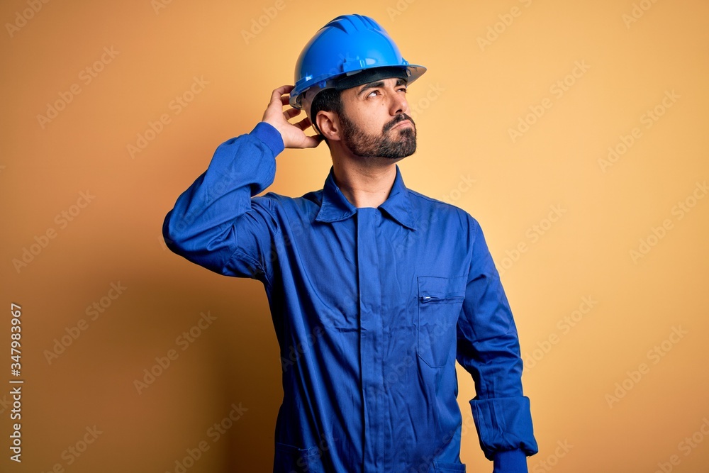 Mechanic man with beard wearing blue uniform and safety helmet over yellow background confuse and wondering about question. Uncertain with doubt, thinking with hand on head. Pensive concept.