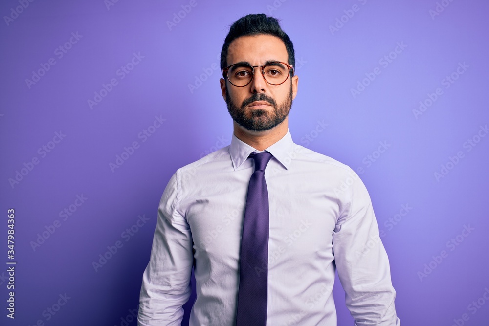 Handsome businessman with beard wearing casual tie and glasses over purple background with serious expression on face. Simple and natural looking at the camera. Handsome businessman with beard wearing casual tie and glasses over purple background with serious expression on face. Simple and natural looking at the camera.