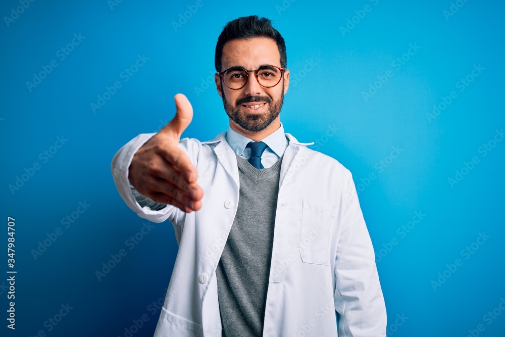 Young handsome doctor man with beard wearing coat and glasses over blue background smiling friendly offering handshake as greeting and welcoming. Successful business. Young handsome doctor man with beard wearing coat and glasses over blue background smiling friendly offering handshake as greeting and welcoming. Successful business.