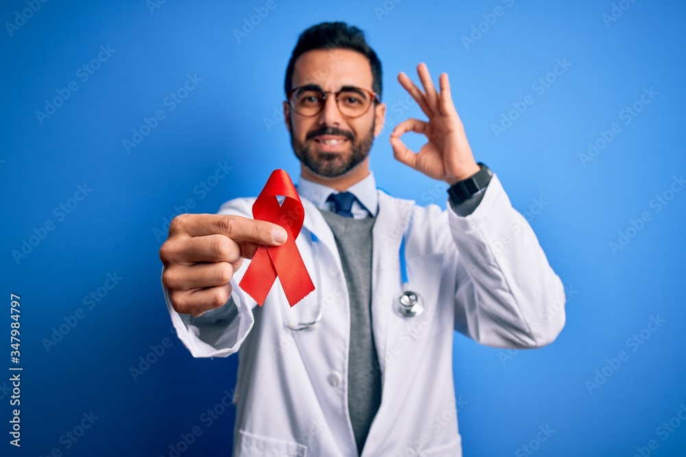 Young handsome doctor man with beard wearing stethoscope holding red hiv ribbon doing ok sign with fingers, excellent symbol Young handsome doctor man with beard wearing stethoscope holding red hiv ribbon doing ok sign with fingers, excellent symbol