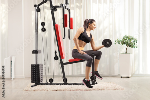 Young female exercising with a dumbbell and sitting on a fitness machine at home