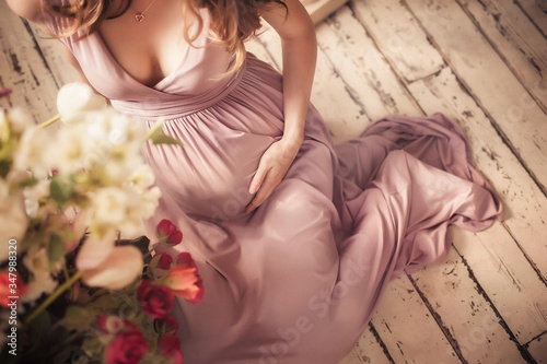 pregnant young woman, in front of a window with magnificent lighting, holds her big belly, in a dress, mother's day, hands on her stomach.