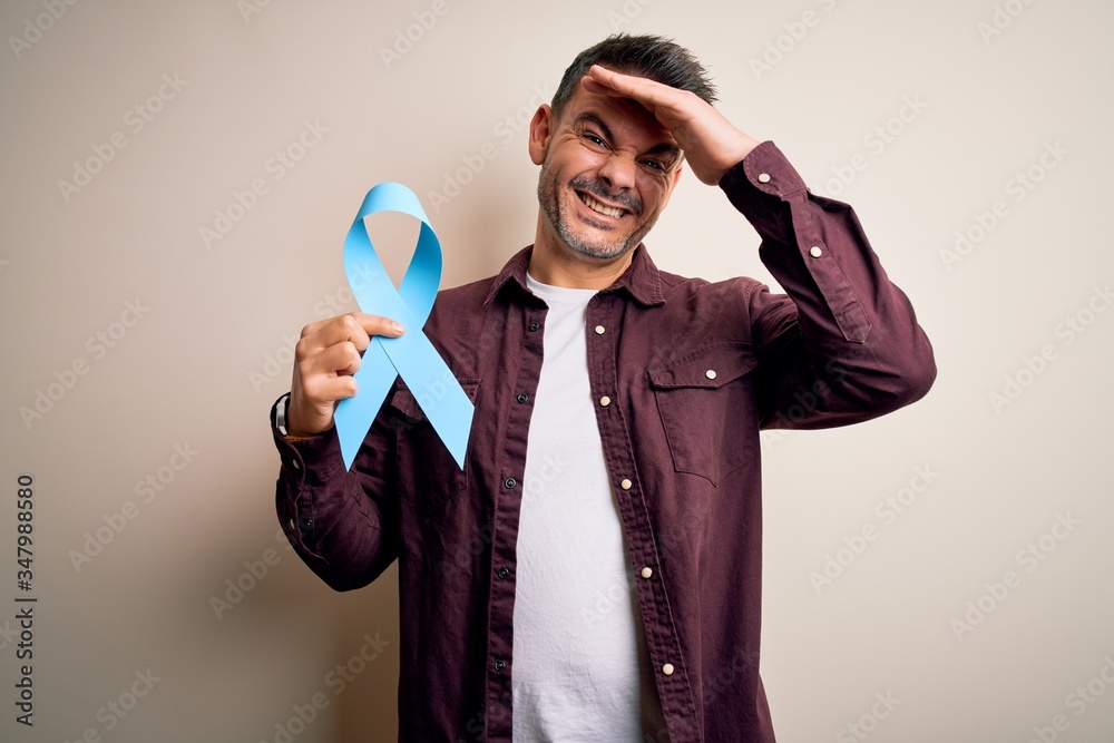 Young handsome man holding blue cancer ribbon over isolated white background stressed with hand on head, shocked with shame and surprise face, angry and frustrated. Fear and upset for mistake. Young handsome man holding blue cancer ribbon over isolated white background stressed with hand on head, shocked with shame and surprise face, angry and frustrated. Fear and upset for mistake.
