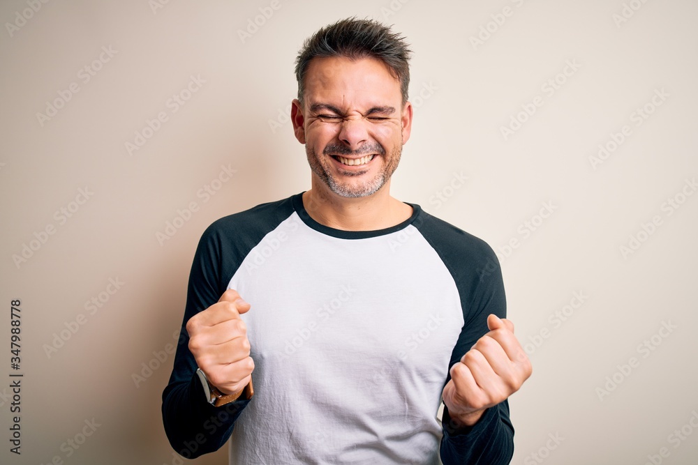 Young handsome man wearing casual t-shirt standing over isolated white background very happy and excited doing winner gesture with arms raised, smiling and screaming for success. Celebration concept. Young handsome man wearing casual t-shirt standing over isolated white background very happy and excited doing winner gesture with arms raised, smiling and screaming for success. Celebration concept.