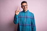 Young handsome man wearing casual shirt and glasses standing over isolated pink background showing and pointing up with finger number one while smiling confident and happy.