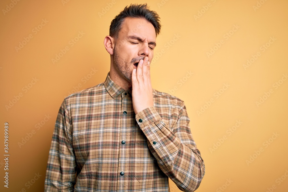 Young handsome man wearing casual shirt standing over isolated yellow background bored yawning tired covering mouth with hand. Restless and sleepiness. Young handsome man wearing casual shirt standing over isolated yellow background bored yawning tired covering mouth with hand. Restless and sleepiness.