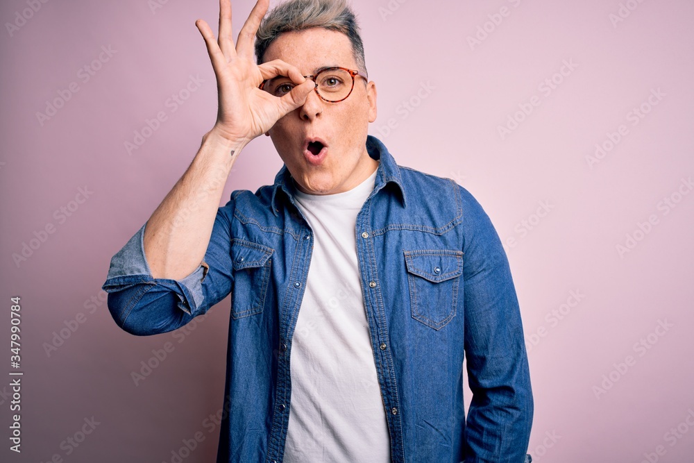 Young handsome modern man wearing glasses and denim jacket over pink isolated background doing ok gesture shocked with surprised face, eye looking through fingers. Unbelieving expression.