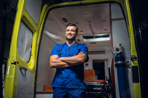 Young paramedic in a blue uniform standing and smiling in front of an an ambulance car
