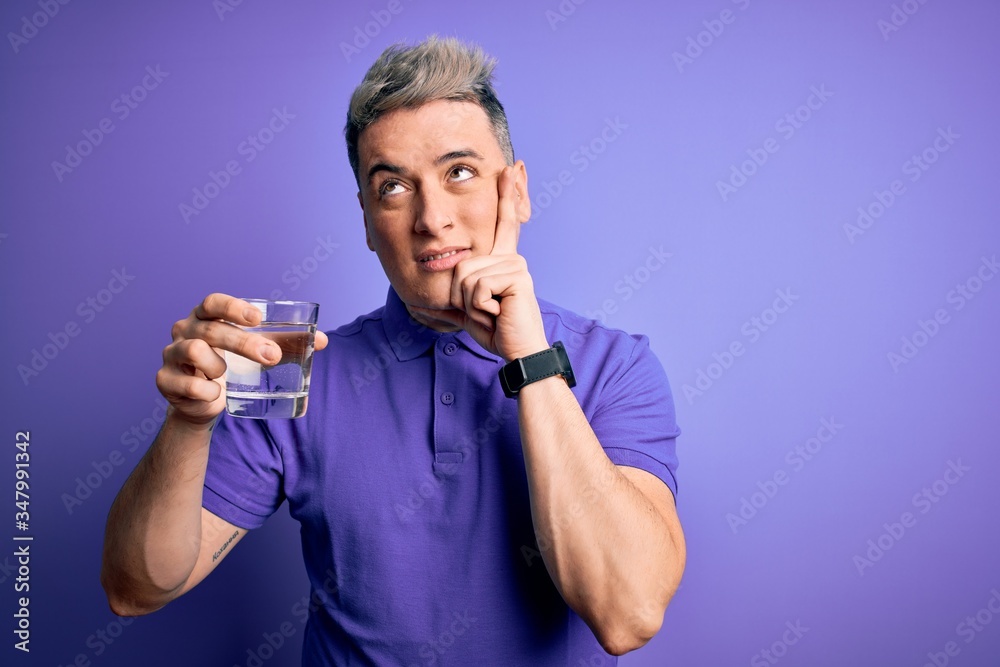 Young modern man drinking a glass of fresh water over purple isolated background serious face thinking about question, very confused idea Young modern man drinking a glass of fresh water over purple isolated background serious face thinking about question, very confused idea