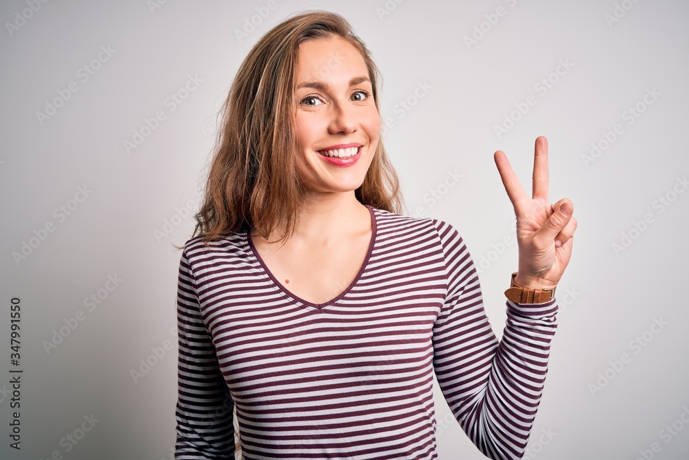 Young beautiful blonde woman wearing casual striped t-shirt over isolated white background smiling with happy face winking at the camera doing victory sign. Number two. Young beautiful blonde woman wearing casual striped t-shirt over isolated white background smiling with happy face winking at the camera doing victory sign. Number two.