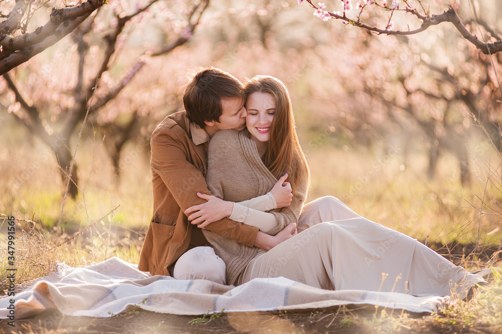 A young man in a brown light jacket gently embraces a beautiful girl with long hair in the back - a couple sitting in nature in a flowering garden. selective front and back focus A young man in a brown light jacket gently embraces a beautiful girl with long hair in the back - a couple sitting in nature in a flowering garden. selective front and back focus