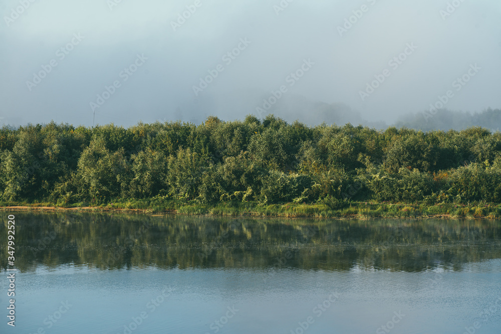 Early morning l Spring landscape with a full flowing river in the foreground and a green Bank overgrown with bushes and trees in the background. The distant plan is drowned in a thick morning fog