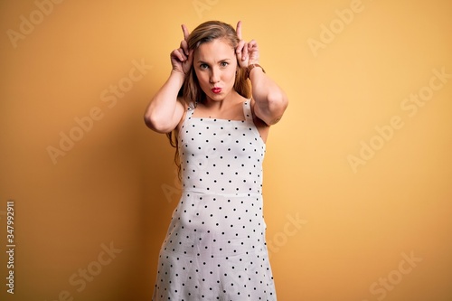 Young beautiful blonde woman on vacation wearing summer dress over yellow background doing funny gesture with finger over head as bull horns