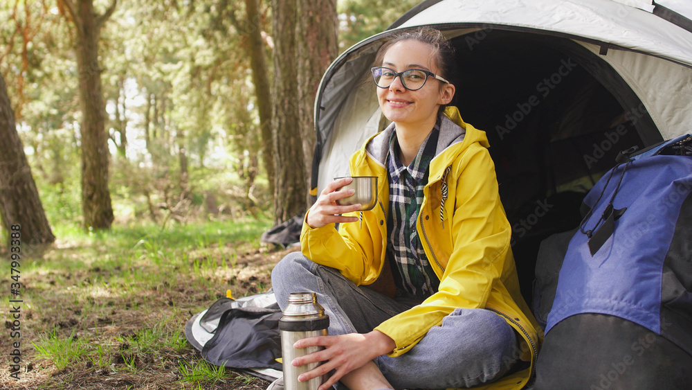 A smiling woman sits near a tent and drinking beverage from thermos. Camping in the forest A smiling woman sits near a tent and drinking beverage from thermos. Camping in the forest