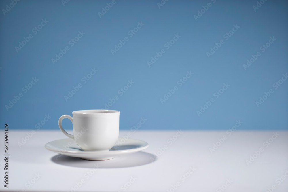 White cup of morning espresso on a stone surface of a table with reflections opposite blue background. White cup of morning espresso on a stone surface of a table with reflections opposite blue background.