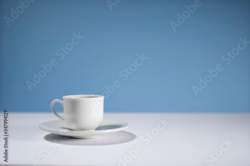 White cup of morning espresso on a stone surface of a table with reflections opposite blue background.