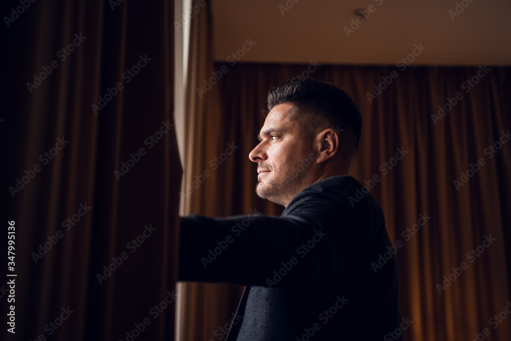 A young businessman in a suit parting the curtains in his hotel A young businessman in a suit parting the curtains in his hotel