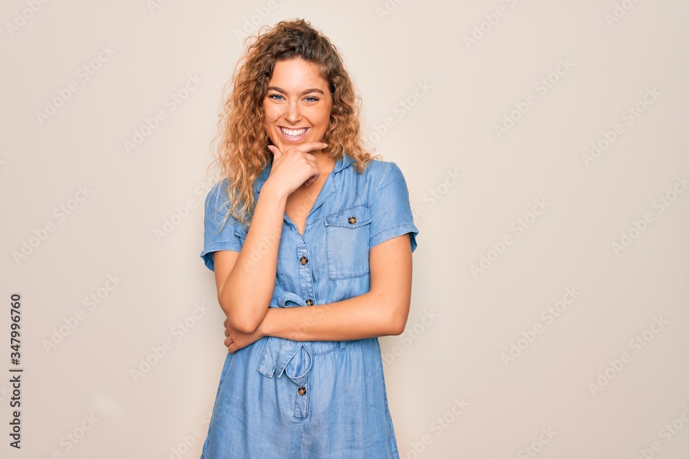 Young beautiful woman with blue eyes wearing casual denim dress over white background looking confident at the camera smiling with crossed arms and hand raised on chin. Thinking positive. Young beautiful woman with blue eyes wearing casual denim dress over white background looking confident at the camera smiling with crossed arms and hand raised on chin. Thinking positive.